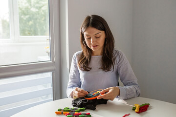 Woman cross-stitches, sitting by window. Hobby of young adult girl.