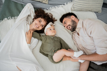 Fototapeta premium top view of positive parents with infant girl lying on bed at home.