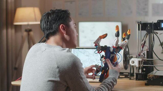 Side View Of Asian Male With 3D Printing Checking And Comparing A Cyborg Hand To The Pictures On A Laptop At Home

