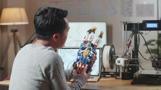 Side View Of Asian Male With 3D Printing Checking A Cyborg Hand While Designing It On A Laptop At Home
