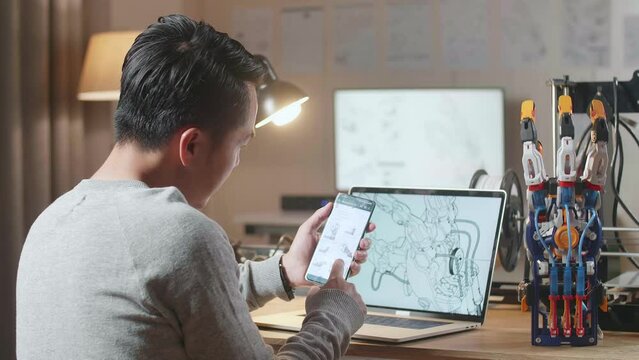 Side View Of Asian Male With 3D Printing Looking At The Pictures On Smartphone And Thinking While Designing A Cyborg Hand On A Laptop At Home

