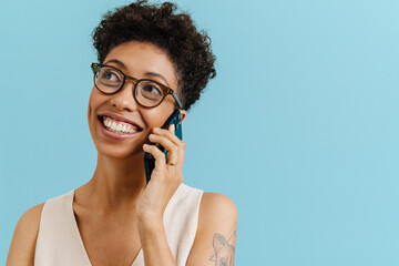 Young curly woman in eyeglasses smiling and talking on cellphone