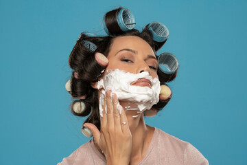 attractive young woman with hair curlers rollers applyes shaving foam on her face on a blue...