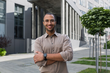 Young successful businessman smiling and looking at camera with crossed arms, man outside office building wearing shirt and glasses, startup investor. - obrazy, fototapety, plakaty