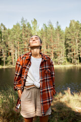Pretty woman in red checkered shirt standing with closed eyes on background with river and forest, inhaling fresh air, enjoying moment of relaxation, listening to tranquility of wild nature
