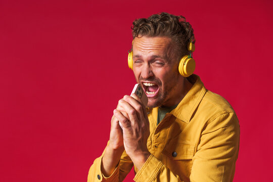 Handsome man 30s antics while pretend singing song using phone and wireless headphones wearing denim yellow jacket isolated on red background. Joyful man sing while listen music