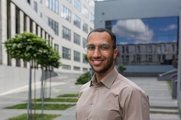 Close-up photo portrait of young entrepreneur wearing glasses, hispanic man smiling and looking at camera, startup entrepreneur outside modern office building.