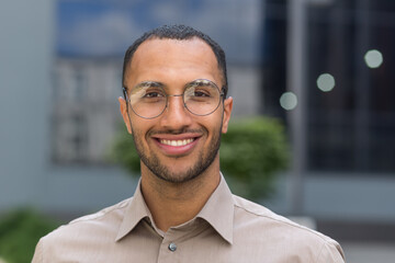 Close-up photo portrait of young entrepreneur wearing glasses, hispanic man smiling and looking at camera, startup entrepreneur outside modern office building.