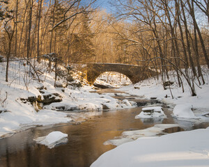 A winter scene with a creek, snow, and an old stone bridge in the forest in early morning light.