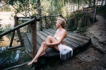 A slender beautiful positive woman with blond hair and a sporty figure, with a stylish hairstyle, in white lingerie, sits on a wooden bridge in a forested area on the hills.