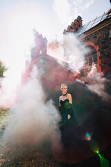 Art work. A slender beautiful woman with blond hair in a black vintage dress and expensive jewelry, stylish makeup and hairstyle, comes out of an ancient castle in fire and smoke.