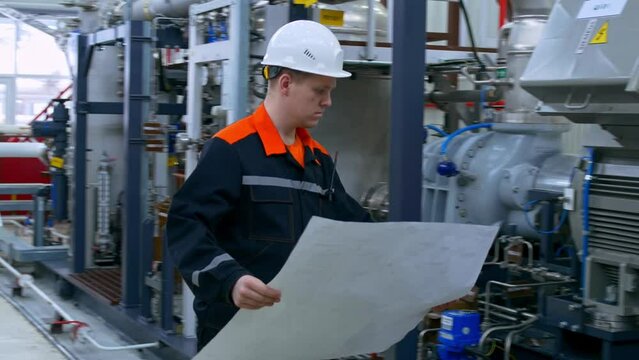 A young industrial engineer in overalls and a white helmet in a factory checks the equipment of a pumping station and checks the diagram in a blueprint along the fuel and lubricant supply pipeline.