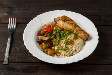 Rice with stewed vegetables in sauce and chicken leg in a white plate on a wooden table.