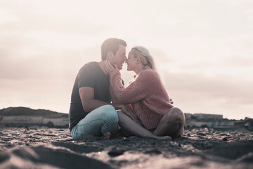 Fototapeta premium Romantic evening with girl and boy together sitting at the beach on the sand kissing in tenderness. Two people male and female in relationship sitting on the sand and enjoying sunset. Man woman kiss