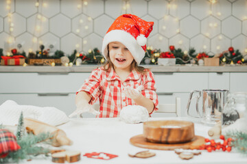 Little dark-haired girl 3 years old in red Christmas cap and checkered shirt prepares dough for gingerbread cookies in white Christmas decorated kitchen with garland lights. Merry Christmas, New Year