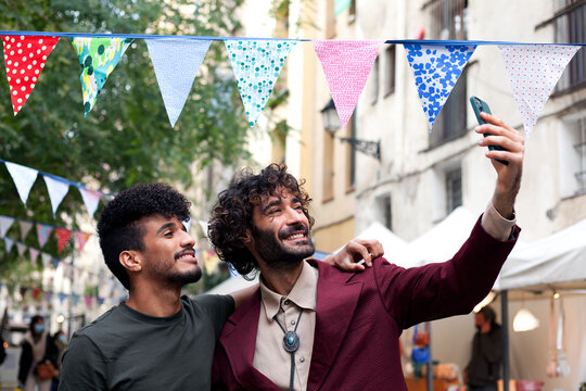 Happy interracial couple of young men taking a selfie while strolling a street adorned with colorful bunting in the background. Two male friends. Urban style concept.