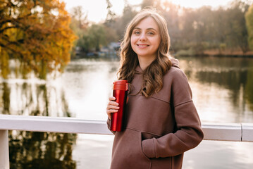 Young woman in hoodie smiling and holding a red thermo mug in an autumn park. Sunny weather. Fall season.