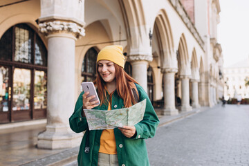 Fototapeta premium Attractive young female tourist is exploring new city. Redhead girl in hat holding a paper map and smartphone on Market Square in Krakow. Traveling Europe in autumn. Active lifestyle concept