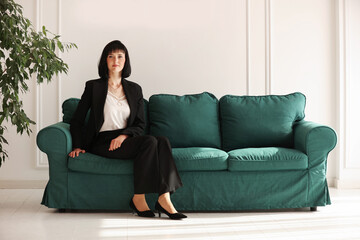 Young businesswoman in suit sitting on the edge of a stylish green sofa in a business lounge with houseplants. Business concept