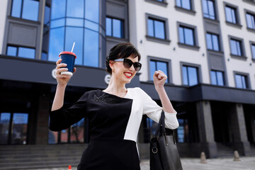 Smiling business woman in sunglasses makes a gesture of the winner standing by business central city street with offices, holding. Successful emotions and gestures. Lifestyle concept