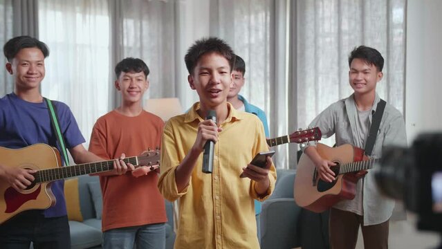 Young Asian Boy, In Front Of His Friends With Musical Instruments, Holding A Microphone And Reading Comments On Smartphone While Live Stream At Home
