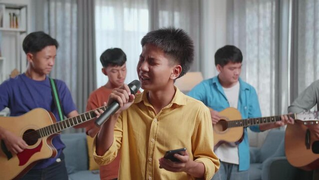 Young Asian Boy Singing And Reading The Lyrics On Smartphone While His Friends Playing Musical Instruments At Home
