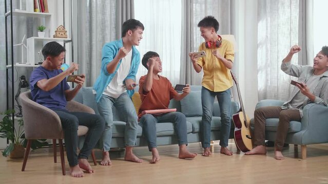 Group Of Young Asian Friends With Musical Instruments Celebrating Winning Game On Smartphones While Sitting On A Sofa At Home
