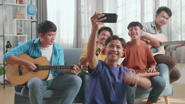 Group Of Young Asian Friends Waving Hand, Singing, And Playing Musical Instruments While Using Smartphone Lives Stream At Home
