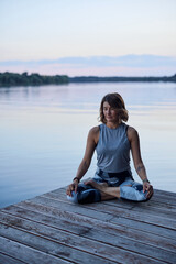Fototapeta na wymiar A calm woman in the lotus pose sits on a dock and practices yoga breathing near the river.