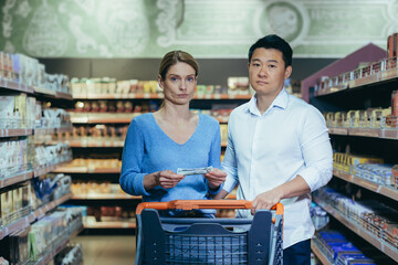 Upset young couple in supermarket, man and woman looking at camera poor have no opportunity and...