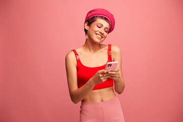 Young woman blogger with colored pink hair and short haircut flipping through the phone screen and typing a message with a smile and surprise in stylish clothes on a pink background