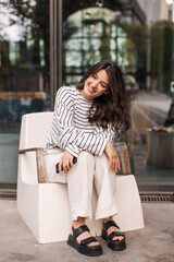 Fototapeta na wymiar Nice young caucasian girl smiling looking away spending time outdoors. Brunette with wavy hair is sitting in chair. Leisure lifestyle and beauty concept.