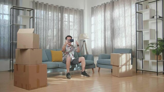 Asian Man With Cardboard Boxes Sitting On The Sofa And Taking Photo By Smartphone After Moving Into A New House 
