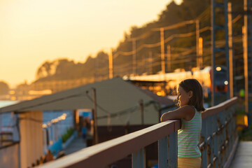 a girl of 8-10 years old stands on a bridge near the railway in the evening at sunset and looks at the sea, a train rides in the background