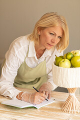 White caucasian blonde matured 
woman makes notes in notebook (planner) and looks at edible fresh bouquet made of fruits apples, rose, lemon, pears, kept on bamboo skewers. Vertical plane