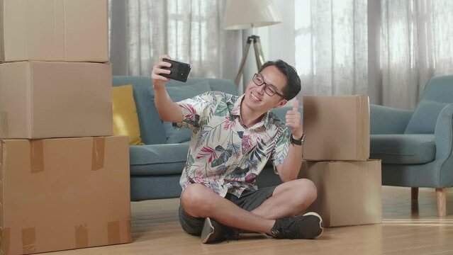 Asian Man With Cardboard Boxes Sitting On The Floor And Taking Photo By Smartphone After Moving Into A New House 
