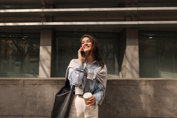Happy young caucasian woman talking on phone outdoors near office. Brunette wears casual clothes, bag and coffee to go. Technology concept