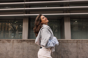 Cheerful young caucasian woman looking away posing standing near city building. Brunette with wavy hair wears classic clothes. Lifestyle concept sincere emotions of people