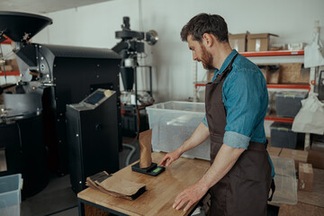 Man barista weighs paper bag with coffee beans on a scale at a coffee factory