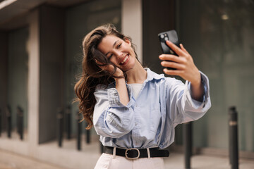 Cute caucasian young girl communicates by video via smartphone standing near business center. Brunette with wavy hair wears blue shirt to street. Cell phone usage concept