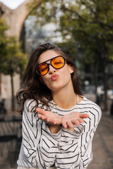 Beautiful young caucasian girl sends air kiss to camera standing outdoors. Brunette with wavy hair holds hands in front of her. Lifestyle, different emotions, leisure concept.