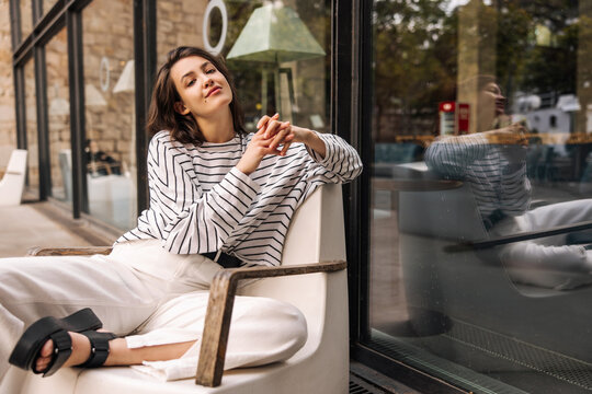 Pretty young caucasian brunette woman posing looking at camera sitting outdoors. Model wears light-colored casual clothes in spring. City life concept