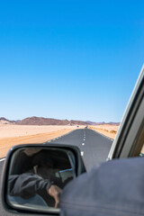 Fototapeta na wymiar Sahara Desert Road trip. Shot from the rear and outside a moving car, while the touareg driver is visible in the mirror but unrecognisable. Blurred blue sky and rocky mountains in Djanet, Algeria.