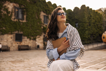 Nice young caucasian woman with textbook for test task sits on campus. Brunette wears casual classic clothes and sunglasses. Modern education, student concept.