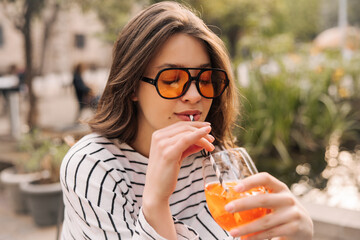 Close-up tender young caucasian girl sipping fruit drink outdoors with her eyes closed. Brunette wears sunglasses in summer day. Leisure lifestyle concept