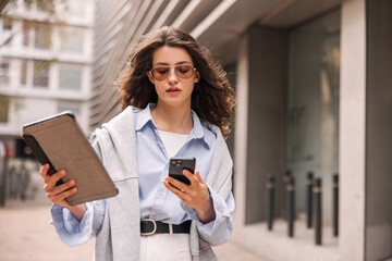 Fototapeta na wymiar Business young caucasian woman uses two wireless devices outdoors. Brunette with flying hair wears sunglasses, shirt, sweatshirt. Management concept