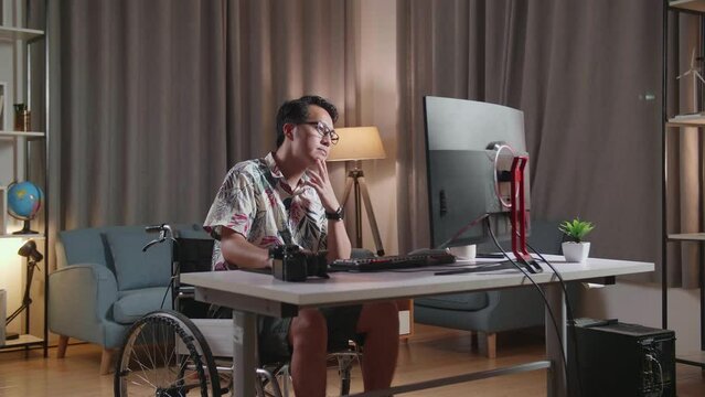 Asian Man In Wheelchair Holding His Chin And Thinking About Something While Editing The Video By A Desktop Next To The Camera At Home
