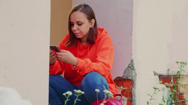 Young woman with short hair in orange hoodie using smartphone, squatting, leaning against wall on street.