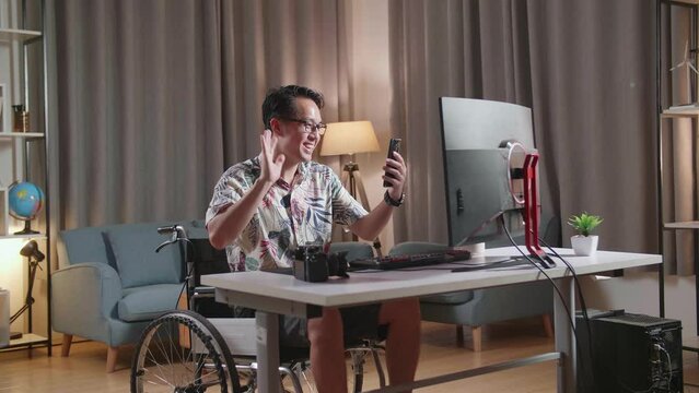 Asian Man In Wheelchair Waving Hand And Having A Video Call On Smartphone While Editing The Video By A Desktop Next To The Camera At Home
