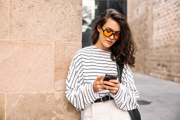 Pretty young caucasian woman chatting with smartphone outdoors. Brunette wears sunglasses, sweatshirt and trousers. Mobile phone usage concept